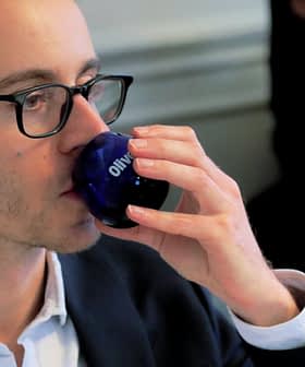 Man wearing glasses drinking from a blue glass with the word 'Olive' on it. - Olive Oil Times