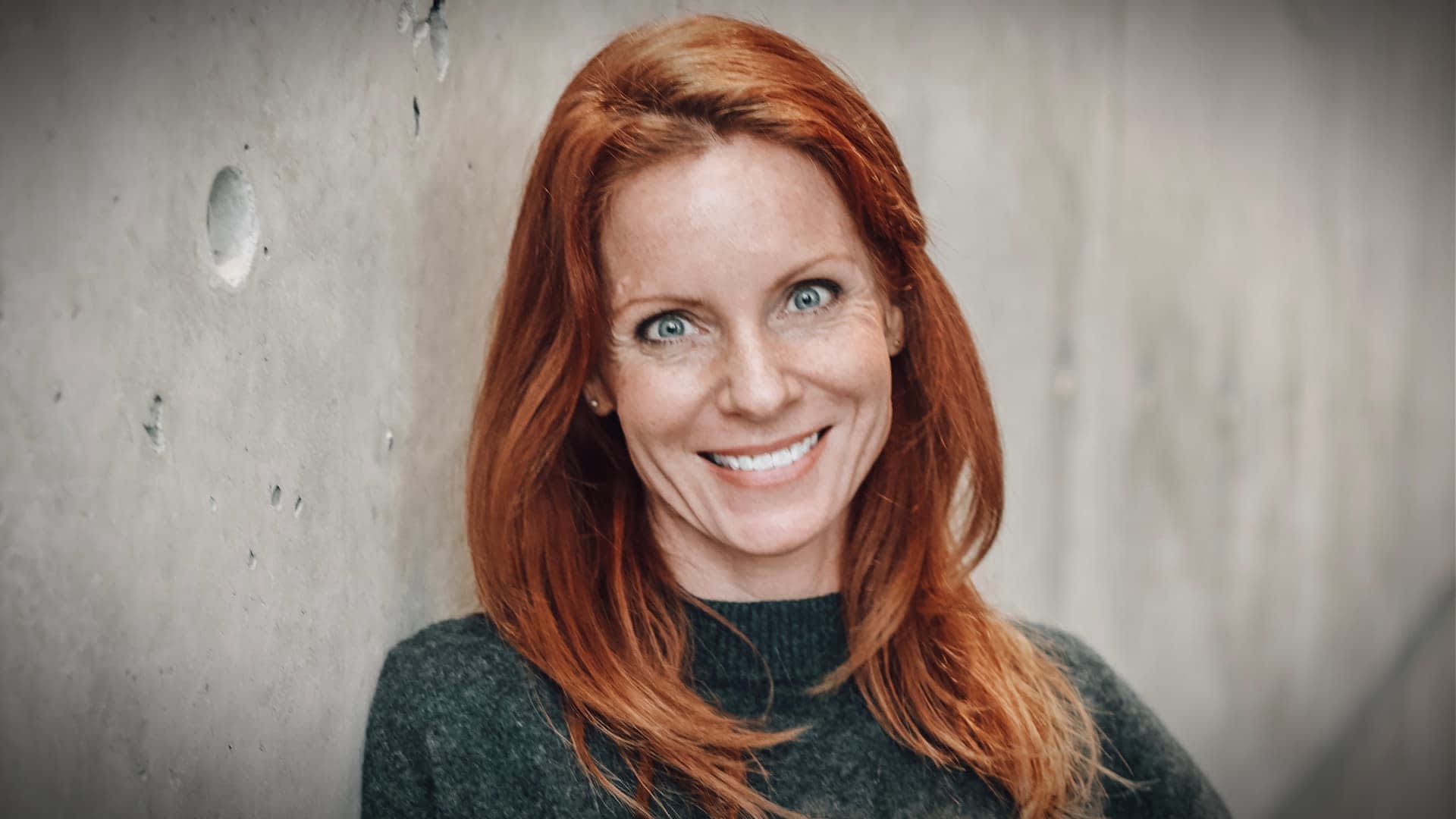 Woman with red hair smiling while leaning against a textured wall. - Olive Oil Times