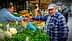 A man in a blue cap interacts with a vendor at a vegetable market filled with various fresh produce. - Olive Oil Times