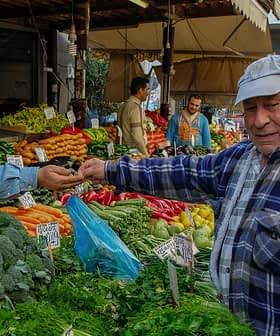 Man in a blue plaid shirt interacting with a vendor at a vegetable market stall. - Olive Oil Times