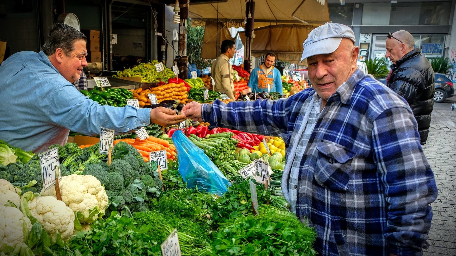 A man in a blue cap interacts with a vendor at a vegetable market filled with various fresh produce. - Olive Oil Times