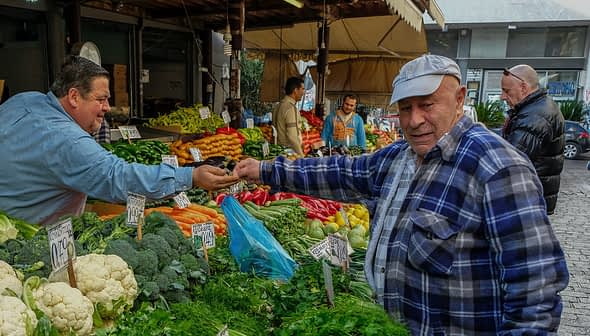 Man in a blue plaid shirt interacting with a vendor at a vegetable market stall. - Olive Oil Times