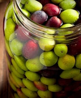 Glass jar filled with a mix of green and black olives on a wooden surface. - Olive Oil Times