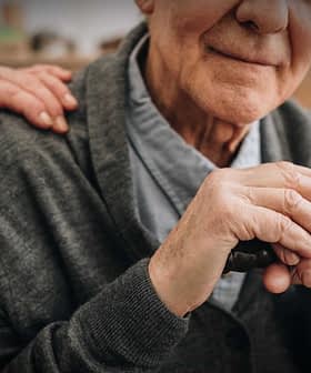 Elderly man with gray hair holding a cane, wearing a gray cardigan, with a hand on his shoulder. - Olive Oil Times