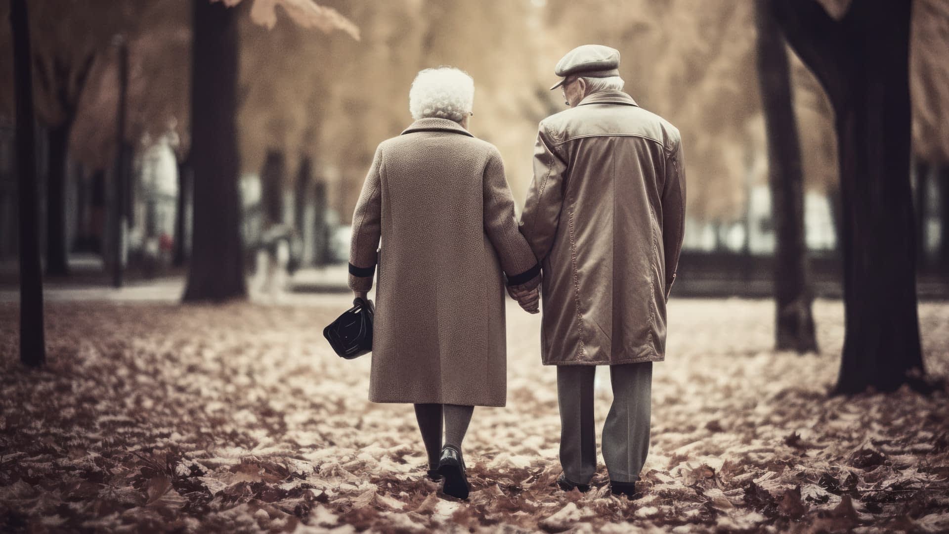 An elderly couple holding hands while walking through a park covered in autumn leaves. - Olive Oil Times