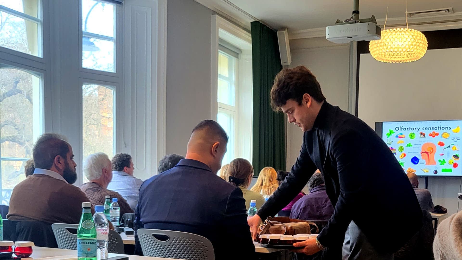 A young man serving food to a seated participant during a presentation on olfactory sensations. - Olive Oil Times