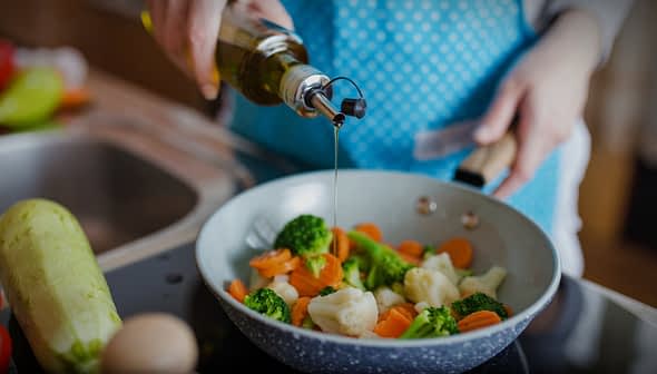 A person in an apron drizzling olive oil over a pan of mixed vegetables including broccoli and carrots. - Olive Oil Times