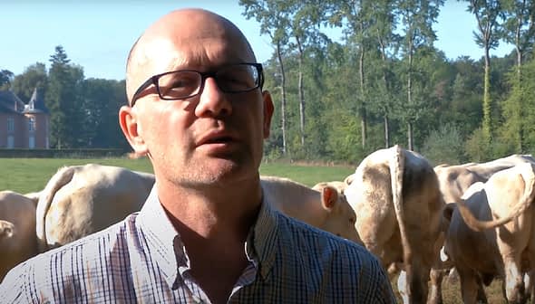 A man wearing glasses and a checked shirt standing in front of a herd of cattle in a field. - Olive Oil Times