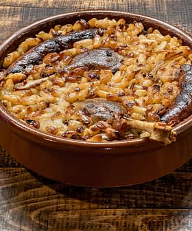 A baked dish containing sausages, onions, and grains in a round ceramic container on a wooden surface. - Olive Oil Times