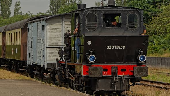 A black steam locomotive with a blue and brown passenger carriage at a train station. - Olive Oil Times