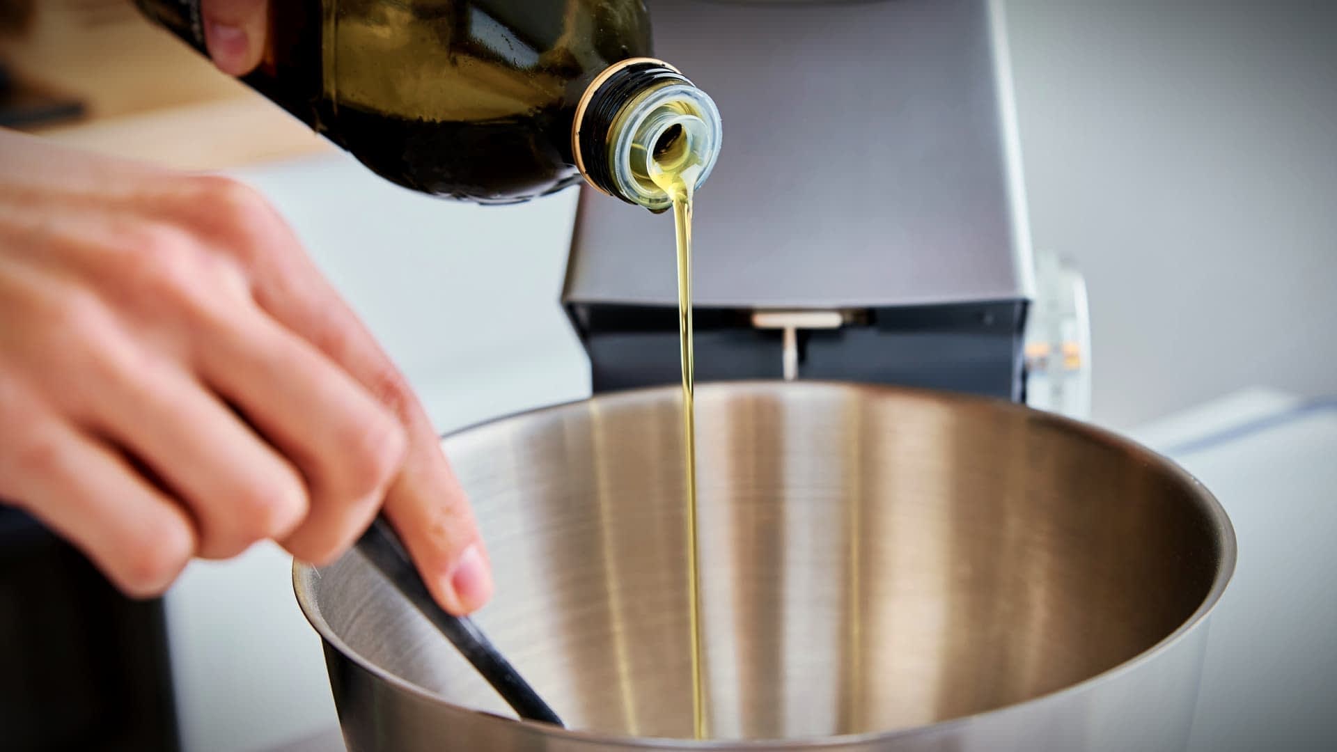 A person pouring olive oil from a bottle into a stainless steel mixing bowl. - Olive Oil Times