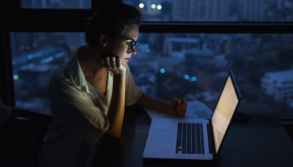 Woman with glasses writing notes while looking at a laptop in a dimly lit environment at night. - Olive Oil Times