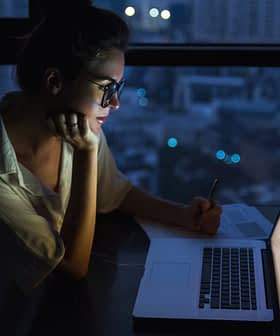 Woman with glasses writing notes while looking at a laptop in a dimly lit environment at night. - Olive Oil Times