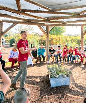 A group of children and adults seated in a circular outdoor learning space with a speaker in the center. - Olive Oil Times