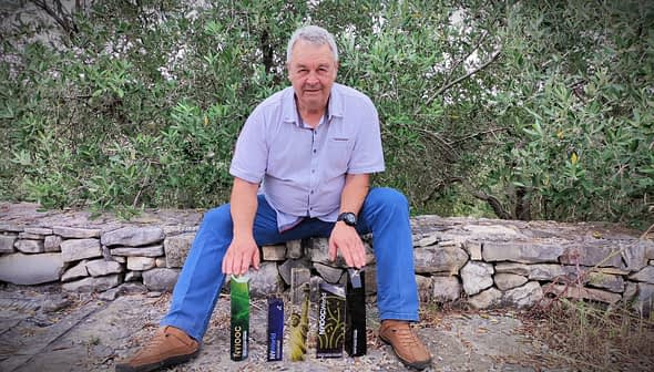 A man seated on a stone wall with various olive oil bottles displayed in front of him. - Olive Oil Times