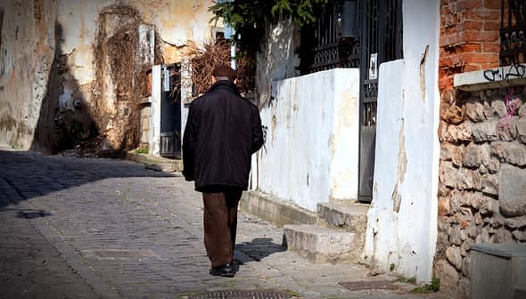An elderly man walking away on a cobblestone street lined with buildings and greenery. - Olive Oil Times