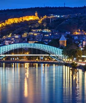 Illuminated bridge over a river with a cityscape and hillside in the background at night. - Olive Oil Times