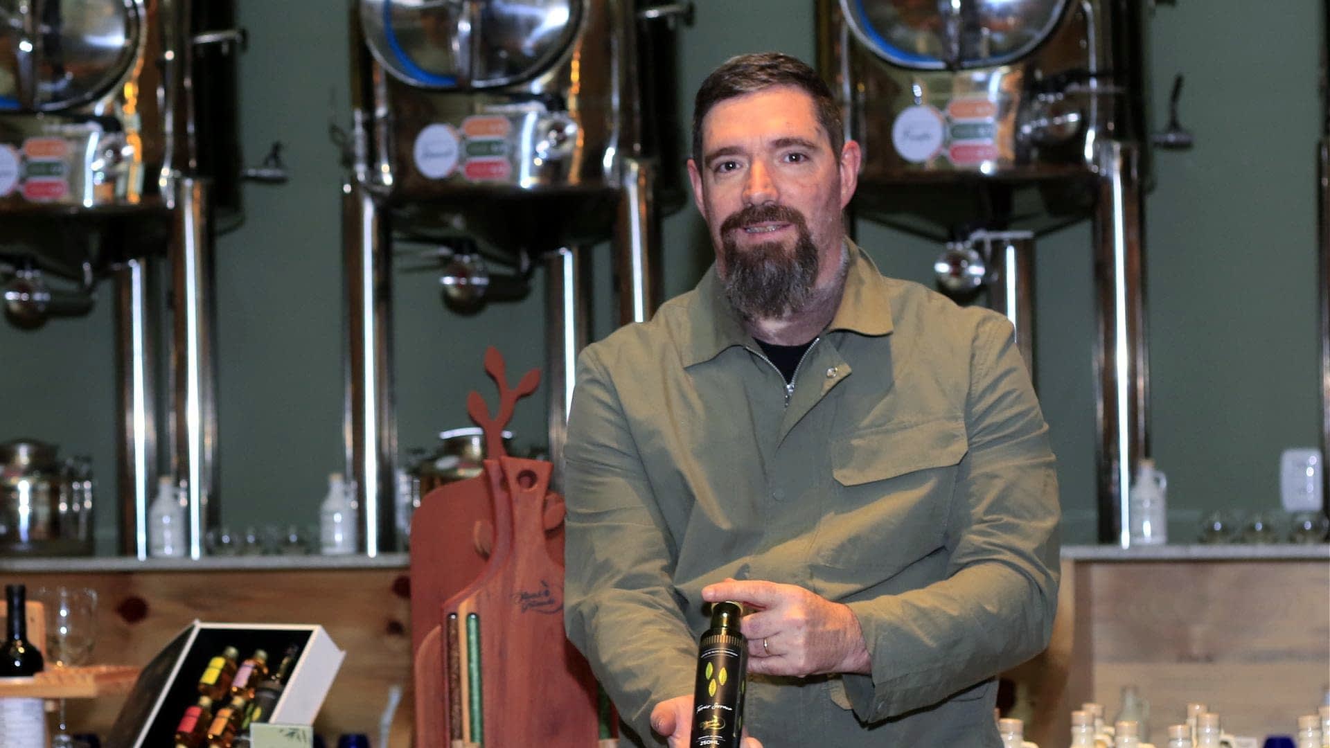 Man with a beard holding a bottle of olive oil in a production facility with metal containers in the background. - Olive Oil Times
