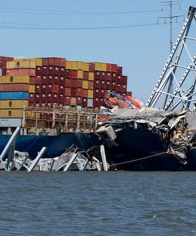 Cargo ship with shipping containers partially submerged and damaged, leaning against a collapsed structure. - Olive Oil Times