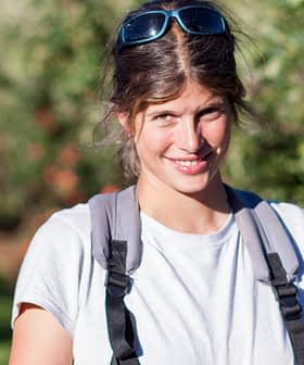 A woman with sunglasses and a backpack smiling while standing in an orchard with trees in the background. - Olive Oil Times