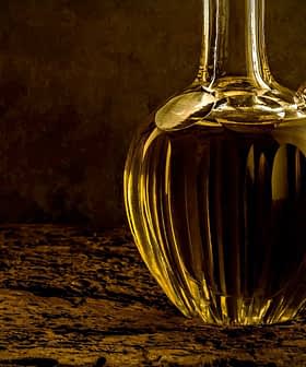 A glass decanter filled with olive oil, featuring a ribbed design and a curved spout. - Olive Oil Times