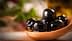 A wooden spoon holding several black olives in olive oil, with a blurred background. - Olive Oil Times