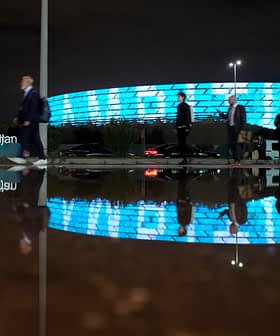 Group of people walking past a modern building with illuminated blue facade at night, reflected in a puddle. - Olive Oil Times