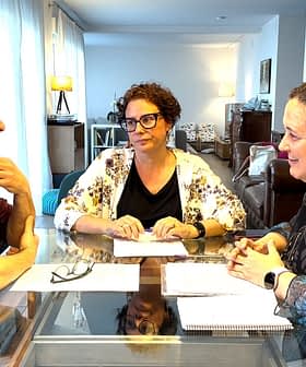 Three individuals seated at a table engaged in a discussion with papers in front of them. - Olive Oil Times