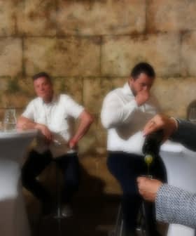 A woman in a gray blazer pouring olive oil while a group of men observes at a tasting event. - Olive Oil Times