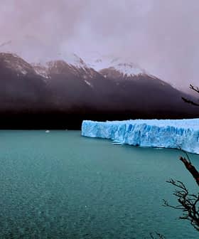 A glacier with a blue ice edge in a body of water, surrounded by mountains and a cloudy sky. - Olive Oil Times