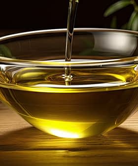 A clear glass bowl filled with olive oil, with olive branches placed nearby on a wooden surface. - Olive Oil Times