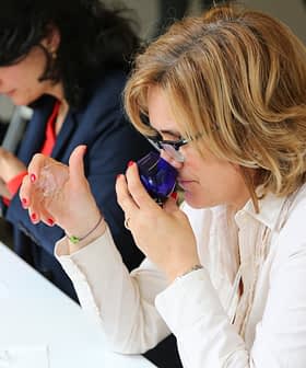 Two women participating in an olive oil tasting session, holding blue glasses and examining the aroma. - Olive Oil Times