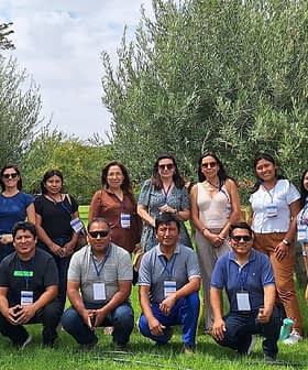 A group of 14 people standing on grass with trees in the background. Some are wearing name tags and sunglasses. The setting appears to be a park or garden.