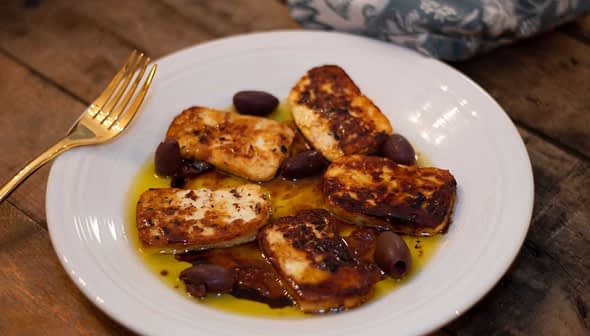 Plate of fried halloumi cheese slices served with black olives and olive oil. - Olive Oil Times