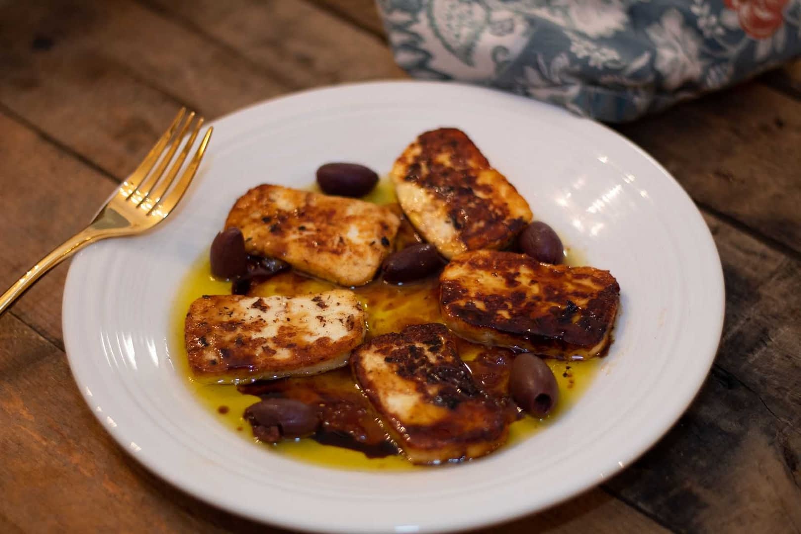 Plate of fried halloumi cheese slices served with black olives and olive oil. - Olive Oil Times