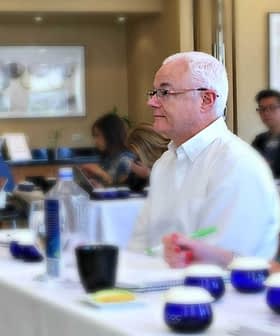 Man with white hair seated at a conference table with participants in the background during a meeting. - Olive Oil Times