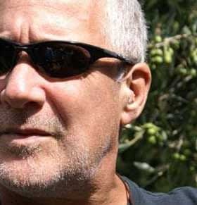 Man with short gray hair wearing sunglasses, with olive trees in the background. - Olive Oil Times