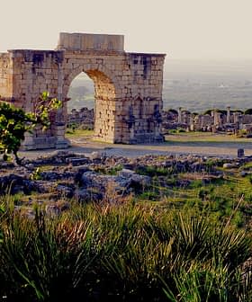 Stone archway structure at an archaeological site with surrounding ruins and greenery. - Olive Oil Times
