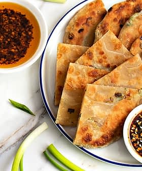 A plate of scallion pancakes arranged with two bowls of dipping sauces and green onions on a marble surface. - Olive Oil Times