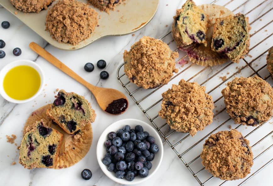 Freshly baked blueberry muffins with a crumb topping on a cooling rack and a plate of blueberries. - Olive Oil Times
