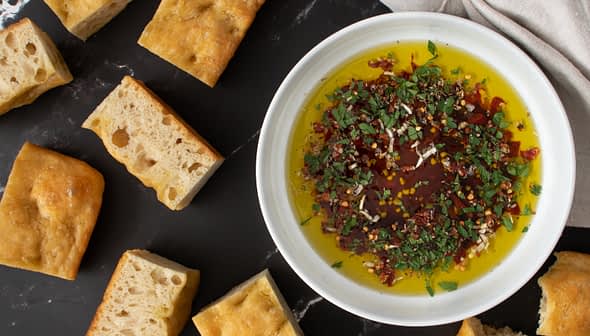 Bowl of olive oil mixed with herbs and spices surrounded by pieces of bread on a dark surface. - Olive Oil Times