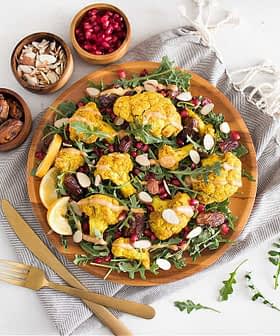 A vibrant salad featuring roasted cauliflower, arugula, nuts, and pomegranate seeds on a wooden plate. - Olive Oil Times