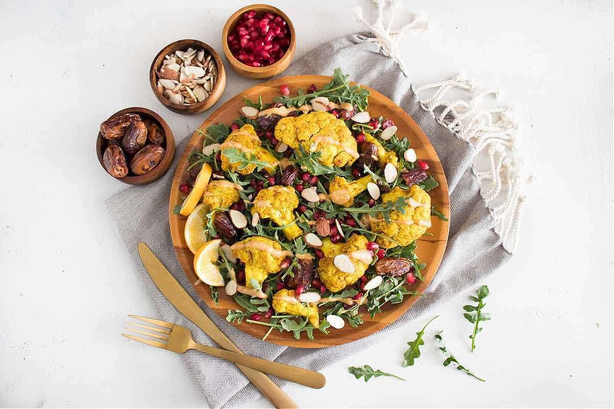 A vibrant salad featuring roasted cauliflower, arugula, nuts, and pomegranate seeds on a wooden plate. - Olive Oil Times