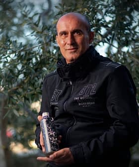 Man in a black jacket holding a bottle while standing among olive trees. - Olive Oil Times