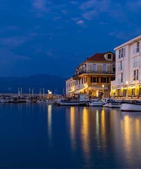 Waterfront scene featuring boats docked near a building with lights reflecting on the water at dusk. - Olive Oil Times