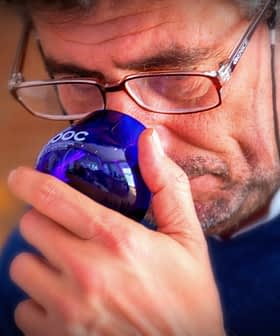 Man with glasses holding a blue glass cup to his nose, appearing to smell it. - Olive Oil Times
