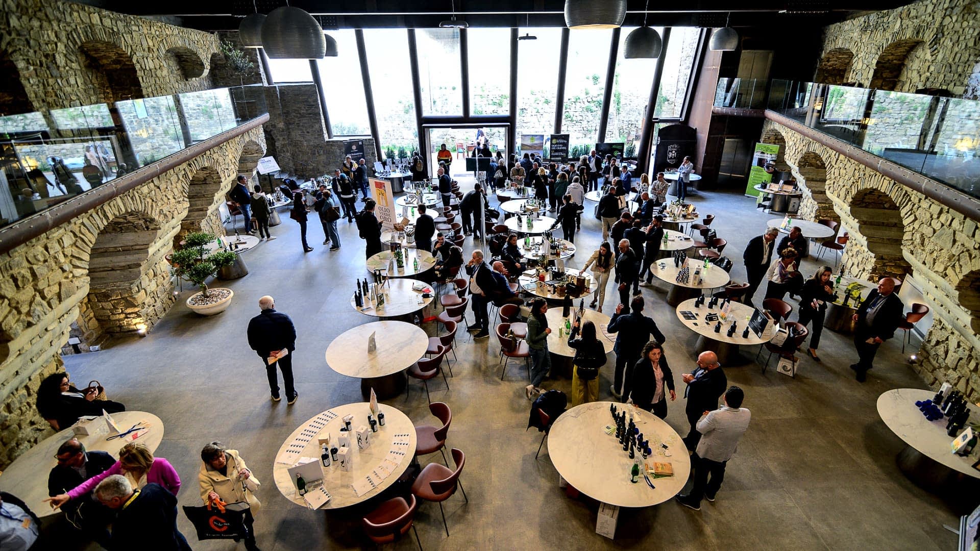 Interior of a spacious event venue featuring round tables and attendees engaged in conversation. - Olive Oil Times