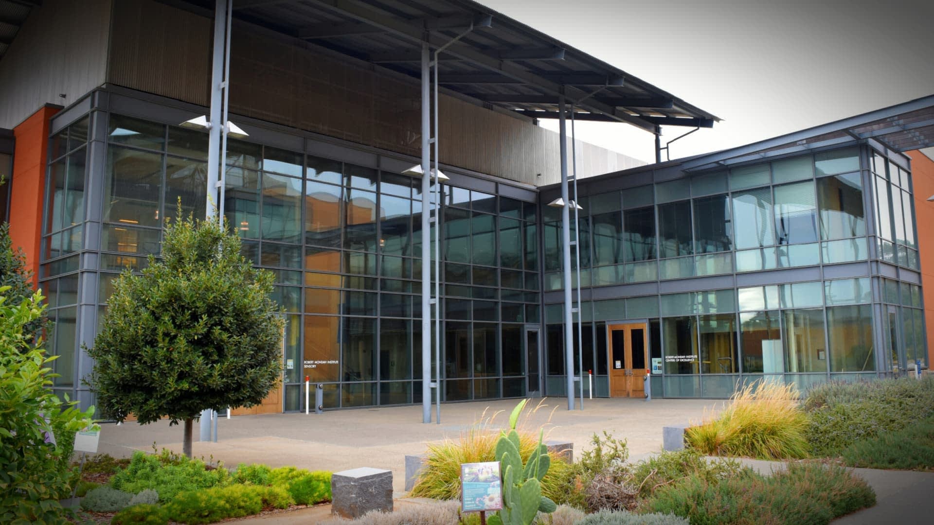 Entrance of a modern building featuring large glass windows and a metal roof structure. - Olive Oil Times