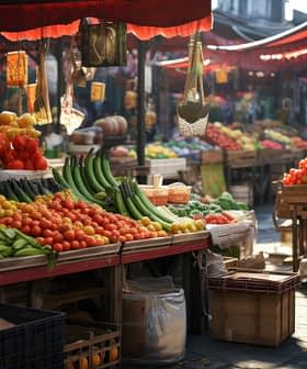Outdoor market with various fruits and vegetables on display under red umbrellas, with vibrant colors and sunlight creating a lively atmosphere.