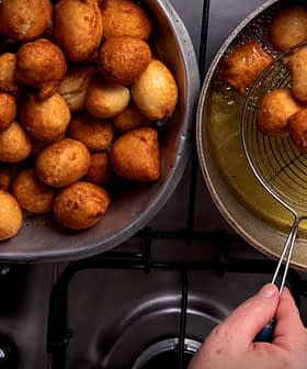A bowl filled with golden-brown fried dough balls next to a frying pan with more dough balls being drained. - Olive Oil Times